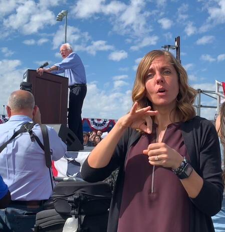 Interpreter outdoors signing vote. Bernie Sanders in the background at a podium.